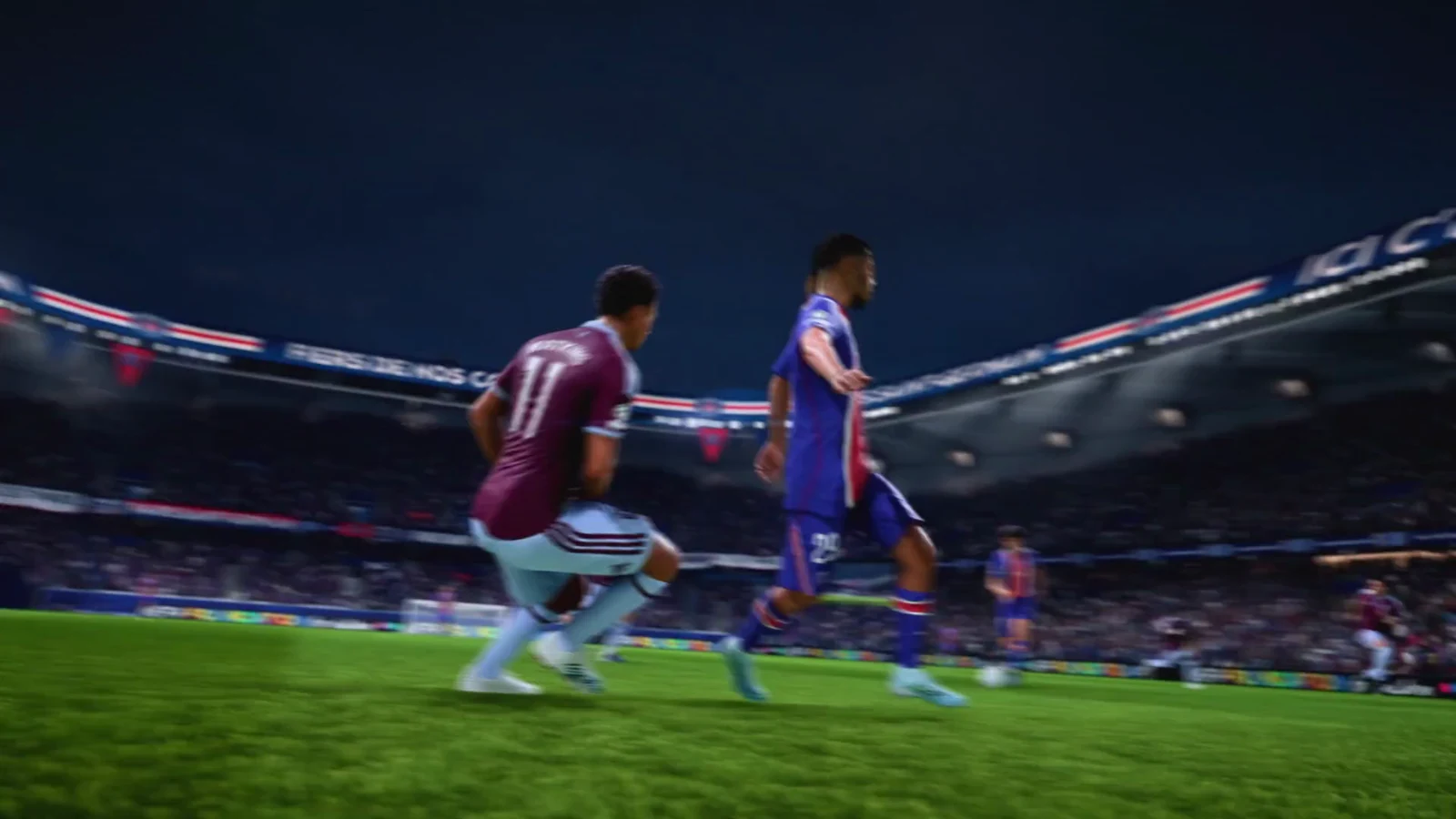 Maroon and blue footballers chase the ball across a crowded stadium pitch during a nighttime match.