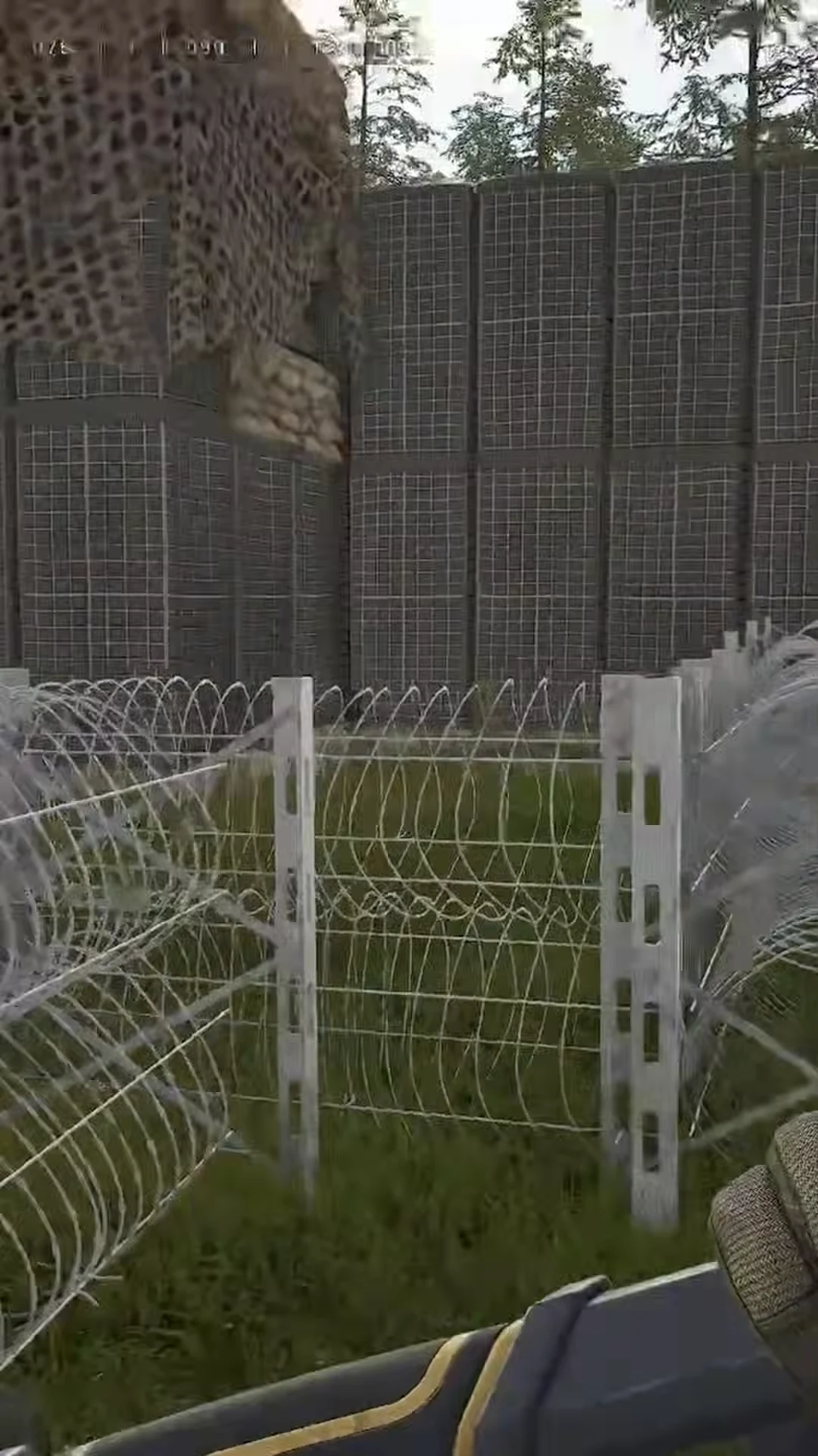 Player walking the outside of a FOB perimeter made of wire fencing, razor wire, and a fortified wall
