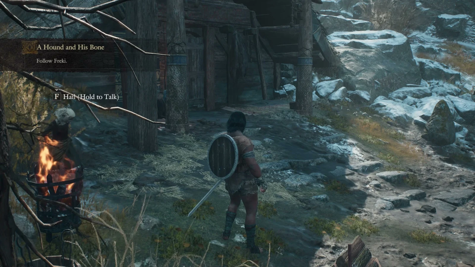 Player with a round shield on their back stands by a wooden lodge and a burning brazier, with a quest banner and talk prompt visible.