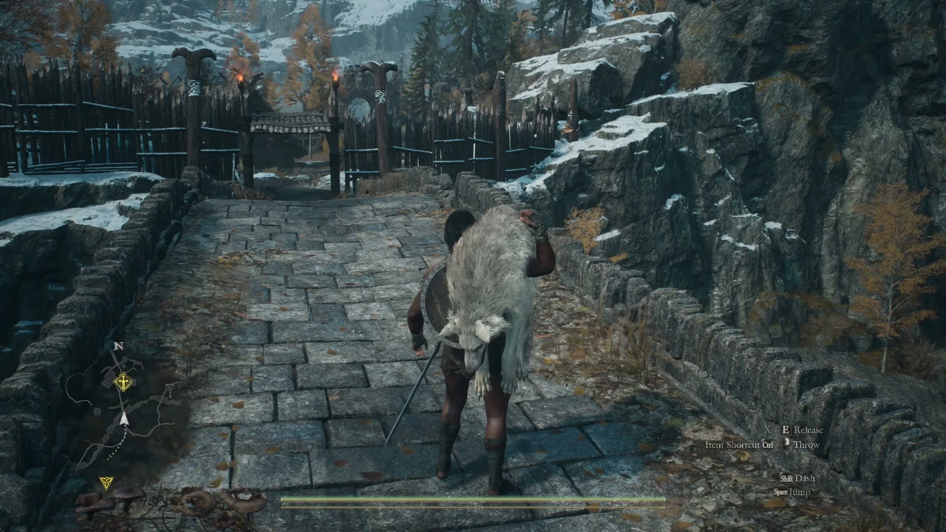 The Arisen carries the white pup Skoll on one shoulder across the stone bridge toward the torch-lit gate in the settlement palisade